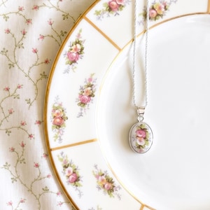 May include: A silver necklace featuring an oval pendant with a floral rose pattern. The pendant displays a cluster of pink and yellow roses on a white background, matching the rose motif on the white and gold rimmed porcelain plate.