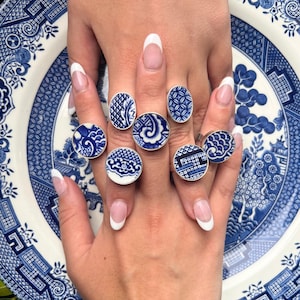 May include: A collection of silver rings featuring blue and white porcelain designs. The rings showcase various patterns, including floral and geometric motifs, reminiscent of classic blue willow china. The rings are displayed on a blue and white patterned plate.