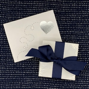 May include: A white gift box with a navy blue ribbon tied in a bow. The box is sitting on a blue and white patterned surface. A white card with a silver heart and a dashed line design is behind the box.