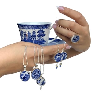 May include: A collection of blue and white jewelry and a teacup with a matching saucer. The jewelry includes a ring, earrings, and three pendant necklaces. The jewelry and teacup feature a classic blue and white Willow pattern.