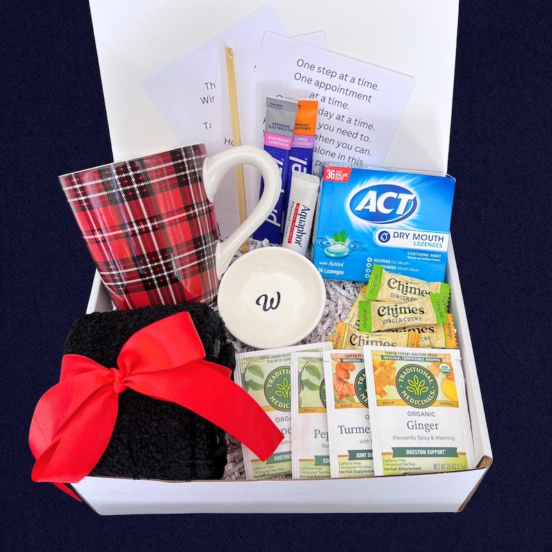 Peut inclure: Une bo&icirc;te cadeau blanche remplie d'articles de soins personnels. Comprend une tasse &agrave; carreaux rouges, un gant de toilette noir avec un n&oelig;ud rouge, un petit bol blanc avec un "w", des sachets de th&eacute;, des pastilles et un baume &agrave; l&egrave;vres. La bo&icirc;te contient une note avec le texte "One step at a time."