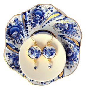 Puede incluir: Pendientes dorados con diseños florales redondos en azul y blanco, adornados con pequeñas perlas y cristales azules. Los pendientes descansan sobre un plato de cerámica blanco, con un diseño floral a juego y ribete dorado.