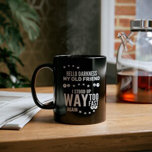 May include: Black coffee mug with white text that reads "Hello darkness my old friend. I stood up way too fast again." The mug has a white outline of two eyes.