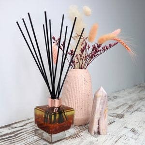 May include: A glass diffuser bottle with black reeds and a pink ceramic vase filled with dried flowers. The diffuser bottle contains a golden liquid with red and orange accents. A pink crystal sits on a white wooden surface.