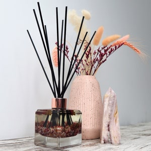 May include: A glass diffuser bottle with black reeds and a pink and white floral arrangement in a pink ceramic vase. The diffuser bottle is filled with a clear liquid and has a rose gold cap. The floral arrangement includes dried flowers and grasses.