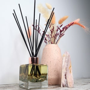 May include: A clear glass diffuser bottle with black reeds and a light brown wooden cap. The bottle is filled with a light green liquid and dried botanicals. The diffuser is sitting on a white surface next to a pink ceramic vase with dried flowers and a pink crystal.