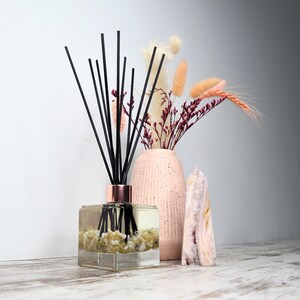 May include: A clear, square diffuser bottle with black reeds and a copper-coloured collar, filled with a light-coloured liquid and small stones. A pink vase with dried flowers and a pink and white crystal are also present.