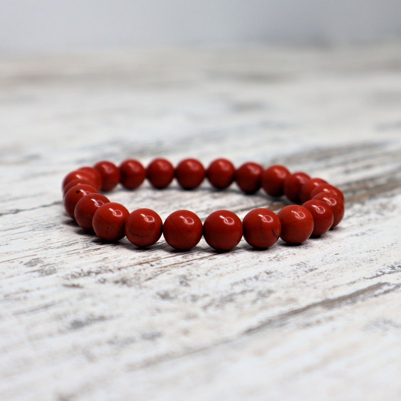 May include: A red jasper beaded bracelet. The bracelet is made of round, polished beads with a deep red color. The beads are strung together to form a circular shape. The bracelet is displayed on a white and gray wooden surface.