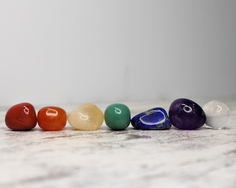 May include: Seven smooth, polished stones in a rainbow of colours, including red, orange, yellow, green, blue, purple, and clear, arranged in a line on a white wooden surface.