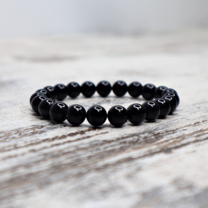 May include: A black beaded bracelet made of round, smooth, polished stones. The bracelet is laid flat on a light wood surface.