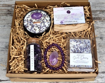 Puede incluir: Una caja de regalo llena de artículos con temática de amatista. Incluye una vela con una piedra púrpura, un frasco de cristales de amatista, una pulsera y ceras perfumadas. La caja también contiene una tarjeta y un palo de madera, todo en papel marrón.