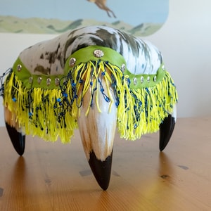 Puede incluir: Un reposapiés con estampado blanco y negro con ribete de flecos verde y amarillo. El reposapiés tiene cuatro patas negras con forma de garras.