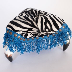 Könnte beinhalten: Ein Fußhocker mit Zebra-Muster, blauem Fransenbesatz und dekorativen Metall-Akzenten. Der Fußhocker hat zwei geschwungene Holzbeine.