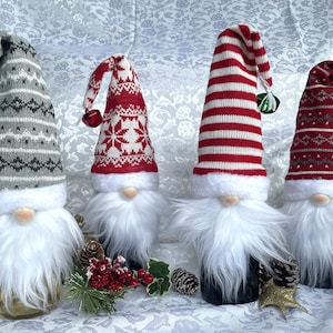 May include: Four gnomes with white beards and different patterned knit hats. The hats are red and white striped, red and white with a snowflake design, gray and white with a geometric design, and red and gray with a geometric design. The gnomes are standing on a white surface with green and red holiday decorations.