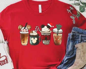 May include: Red t-shirt with a graphic of five Christmas-themed coffee cups with Mickey Mouse ears. The cups are decorated with snowflakes, candy canes, and a Santa hat.