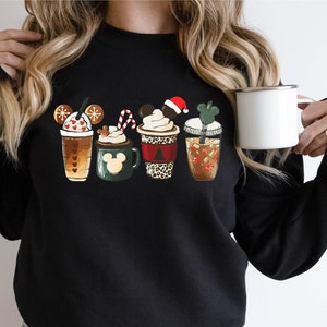 K&ouml;nnte beinhalten: Schwarzes Sweatshirt mit einem Aufdruck von f&uuml;nf verschiedenen weihnachtlichen Kaffeetassen mit Mickey-Maus-Ohren. Die Tassen sind mit Schneeflocken, Zuckerstangen und einer Weihnachtsm&uuml;tze verziert.