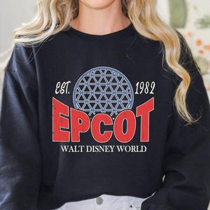May include: Black sweatshirt with a distressed graphic featuring the word "EPCOT" in red, a blue and white geometric sphere, and the text "Walt Disney World" below. The sweatshirt also includes the text "est. 1982".