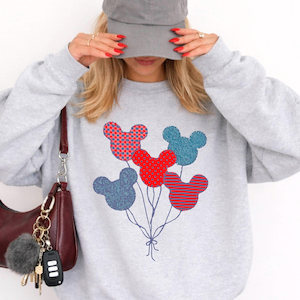May include: A light gray sweatshirt featuring a colorful Mickey Mouse balloon design. The balloons are in red, blue, and pink patterns. A gray baseball cap and a burgundy purse with a keychain are also visible.