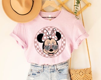 May include: Pink t-shirt featuring a graphic of Minnie Mouse wearing sunglasses and a bow. The graphic is set against a checkered background. The shirt is on a hanger with a straw hat and a woven bag.