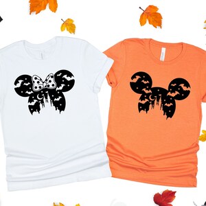 May include: Two t-shirts with a Halloween-themed Mickey Mouse silhouette design. One shirt is white and the other is orange. The design features Mickey Mouse ears with bats and a crescent moon.