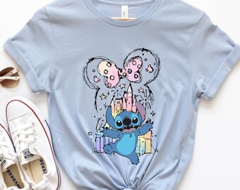 Puede incluir: Camiseta azul claro con un personaje de dibujos animados, un castillo y un diseño inspirado en Minnie Mouse. El gráfico incluye un personaje azul, un castillo rosa y un lazo rosa. La camiseta está colgada en una percha de madera.