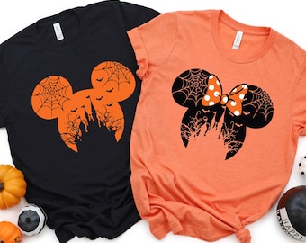 May include: Two Halloween-themed t-shirts. One is black with an orange Mickey Mouse head design featuring a spiderweb, bats, and a haunted castle. The other is orange with a black Mickey Mouse head design with a polka-dot bow.