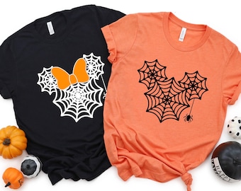 Puede incluir: Dos camisetas con temática de Halloween. Una camiseta es negra con un diseño de cabeza de Mickey Mouse de telaraña blanca. La otra camiseta es naranja con un diseño de cabeza de Mickey Mouse de telaraña negra.