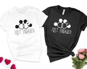 May include: Two t-shirts, one white and one dark grey, each with a graphic of Mickey Mouse ears, two interlocking rings, and a bow, with the text "JUST ENGAGED" in a script font. The shirts are likely for a couple.