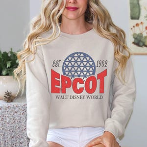 May include: A beige sweatshirt featuring the text "EPCOT" in red, with a blue geometric sphere design above it. The text "EST. 1982" is above the sphere, and "WALT DISNEY WORLD" is below. The sweatshirt has long sleeves.