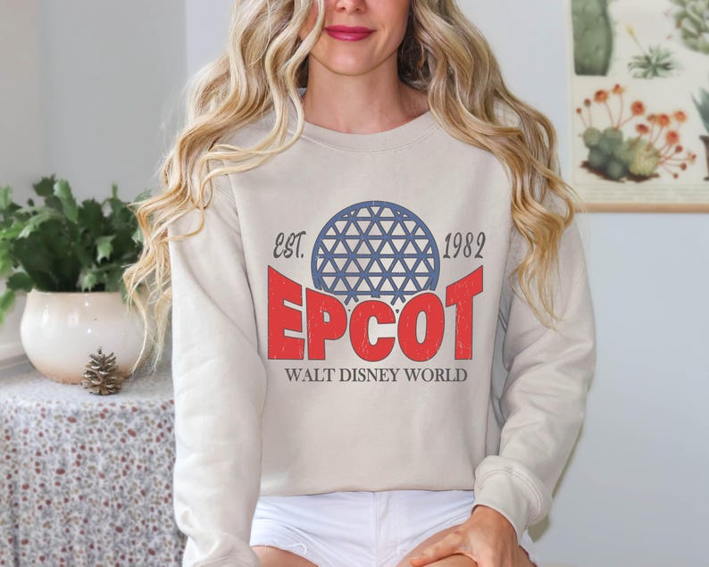 May include: A beige sweatshirt featuring the text "EPCOT" in red, with a blue geometric sphere design above it. The text "EST. 1982" is above the sphere, and "WALT DISNEY WORLD" is below. The sweatshirt has long sleeves.