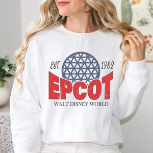 May include: White sweatshirt featuring the text "EPCOT" in red, with a blue geometric sphere design above it. The text "EST. 1982" is above the sphere, and "WALT DISNEY WORLD" is below. A classic, vintage-style design.