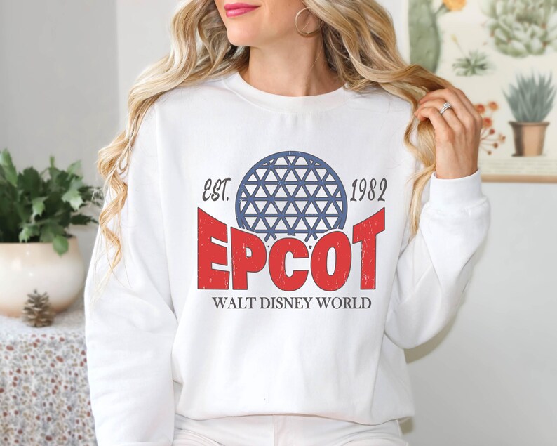 May include: White sweatshirt featuring the text "EPCOT" in red, with a blue geometric sphere design above it. The text "EST. 1982" is above the sphere, and "WALT DISNEY WORLD" is below. A classic, vintage-style design.