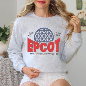 May include: A light gray sweatshirt with a graphic design featuring the word "EPCOT" in red, a blue and white geometric sphere, and the text "EST. 1982" and "WALT DISNEY WORLD". The sweatshirt has long sleeves.