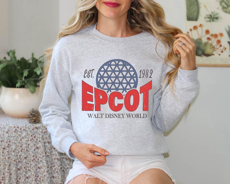May include: A light gray sweatshirt with a graphic design featuring the word "EPCOT" in red, a blue and white geometric sphere, and the text "EST. 1982" and "WALT DISNEY WORLD". The sweatshirt has long sleeves.
