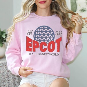 May include: A light pink sweatshirt featuring the text "EPCOT" in red, with a blue geometric sphere design above it. The text "EST. 1982" and "WALT DISNEY WORLD" are also printed on the sweatshirt. The sweatshirt is a crew neck style.