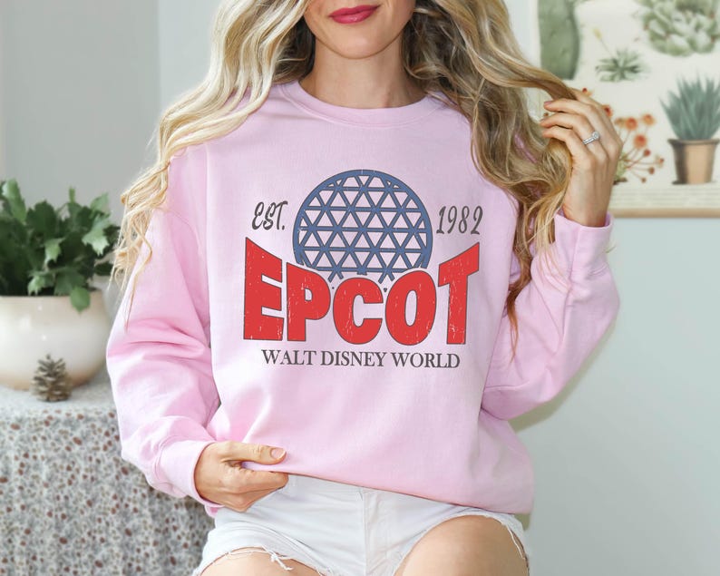 May include: A light pink sweatshirt featuring the text "EPCOT" in red, with a blue geometric sphere design above it. The text "EST. 1982" and "WALT DISNEY WORLD" are also printed on the sweatshirt. The sweatshirt is a crew neck style.