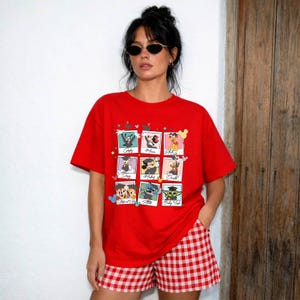 May include: A red t-shirt featuring a grid of nine Disney character portraits. The characters include Goofy, Minnie Mouse, and Donald Duck. The model is wearing red and white checkered shorts.
