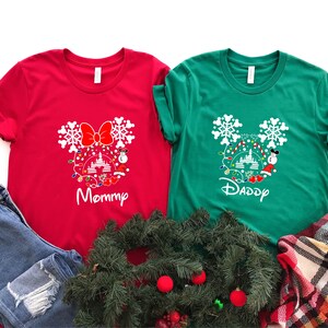 May include: Red and green t-shirts with a white Mickey Mouse silhouette design featuring a Christmas tree, lights, and snowflakes. The text "Mommy" is on the red shirt and "Daddy" is on the green shirt.