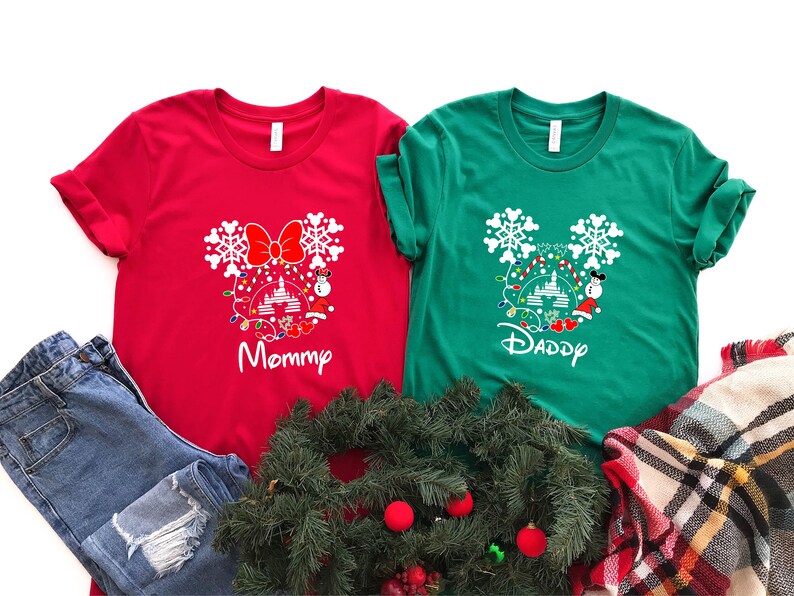 May include: Red and green t-shirts with a white Mickey Mouse silhouette design featuring a Christmas tree, lights, and snowflakes. The text "Mommy" is on the red shirt and "Daddy" is on the green shirt.