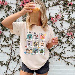 May include: Cream-colored t-shirt featuring a grid of cartoon characters in graduation caps. The characters include Mickey Mouse, Donald Duck, and others. The shirt is worn by a person holding a beverage.