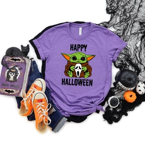 K&ouml;nnte beinhalten: Lilafarbenes T-Shirt mit einer gr&uuml;nen Alienfigur, die eine wei&szlig;e Geistermaske h&auml;lt. Auf dem Shirt steht "Happy Halloween".