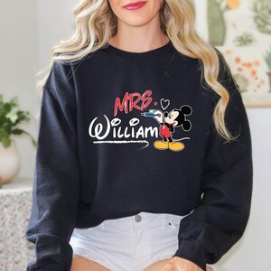 May include: Black sweatshirt with the text "MRS. (William)" in white with a red heart and a cartoon Mickey Mouse holding a pencil.