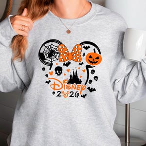May include: A light gray crewneck sweatshirt featuring a black and orange graphic of Mickey Mouse ears. The design includes a spider web, a polka dot bow, a jack-o'-lantern, a castle silhouette, skulls, bats, and the text Disney 2026.