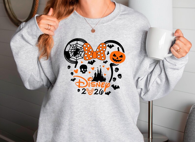 May include: A light gray crewneck sweatshirt featuring a black and orange graphic of Mickey Mouse ears. The design includes a spider web, a polka dot bow, a jack-o'-lantern, a castle silhouette, skulls, bats, and the text Disney 2026.