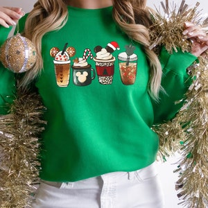 K&ouml;nnte beinhalten: Gr&uuml;ner Weihnachtspullover mit einem Motiv von vier festlichen Kaffeetassen mit Mickey-Maus-Ohren. Die Tassen sind mit Schneeflocken, Zuckerstangen und anderen festlichen Details verziert.