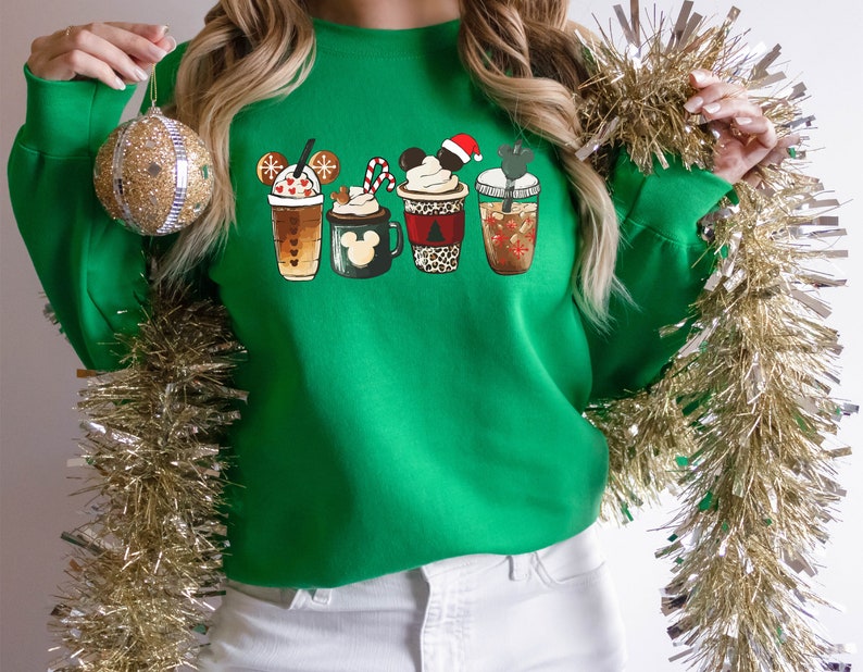 K&ouml;nnte beinhalten: Gr&uuml;ner Weihnachtspullover mit einem Motiv von vier festlichen Kaffeetassen mit Mickey-Maus-Ohren. Die Tassen sind mit Schneeflocken, Zuckerstangen und anderen festlichen Details verziert.