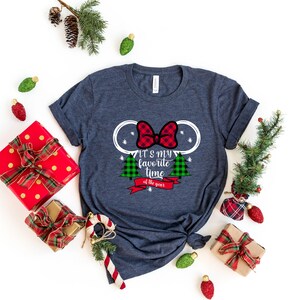 Puede incluir: Una camiseta azul marino con un contorno blanco de las orejas de Mickey Mouse con un lazo de cuadros rojos y negros. La camiseta tiene el texto "It's my favorite time of the year" con un &aacute;rbol de Navidad de cuadros verdes y rojos a cada lado.