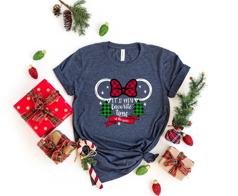 Puede incluir: Una camiseta azul marino con un contorno blanco de las orejas de Mickey Mouse con un lazo de cuadros rojos y negros. La camiseta tiene el texto "It's my favorite time of the year" con un &aacute;rbol de Navidad de cuadros verdes y rojos a cada lado.