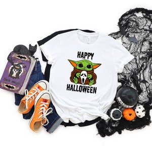 K&ouml;nnte beinhalten: Wei&szlig;es T-Shirt mit einer gr&uuml;nen Baby-Yoda-Figur, die eine wei&szlig;e Geistermaske h&auml;lt. Der Text "HAPPY HALLOWEEN" ist in Schwarz unter der Figur gedruckt.