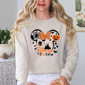 May include: A beige long-sleeve sweatshirt featuring a black and orange graphic of a Mickey Mouse silhouette filled with a spider web, a polka dot bow, a jack-o'-lantern, a castle, skulls, and bats, with the text Disney 2020 printed below.